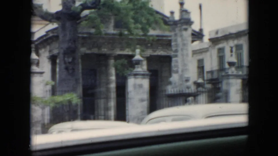 1956 Cuba home-movie frame of a roadside masonry entrance, tree canopy, fence, and car roof viewed from inside a vehicle.