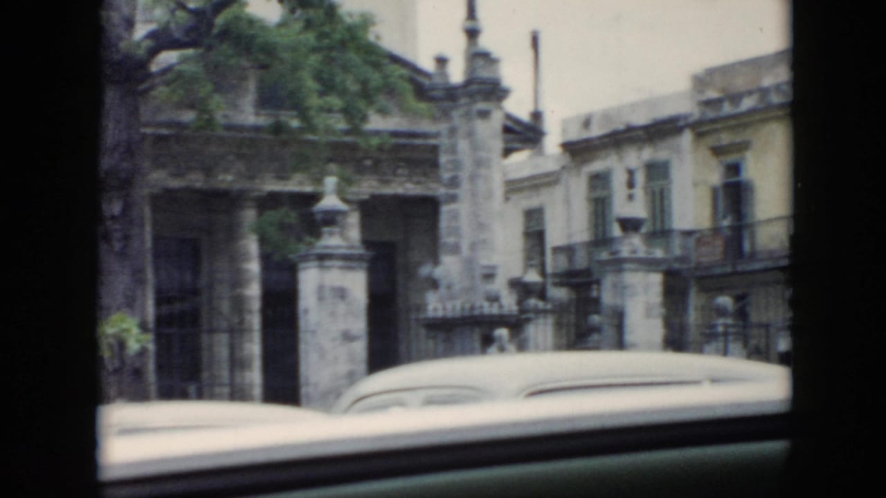 1956 Cuba home-movie frame looking through a car window at a gated stone facade, ornate lamps, trees, and a rounded vintage car roof.
