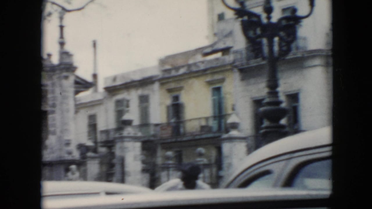 1956 Cuba street view through a car window showing pale buildings, iron balconies, a vintage car, and distant people near the sidewalk.