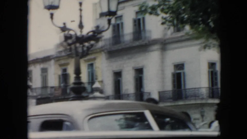 1956 Cuba frame with a soft 8mm view of balconied buildings, an ornate street lamp, and a rounded vintage automobile below.