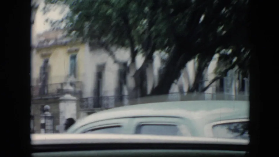 1956 Cuba home-movie frame showing trees, a gated building frontage, pale facades, and a light-colored vintage car from a moving vehicle.