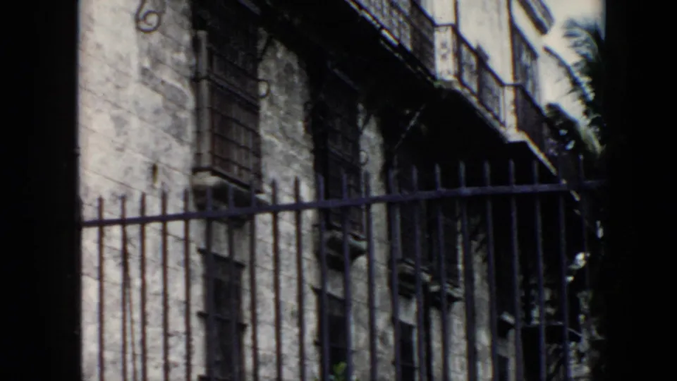 1956 Cuba 8mm frame showing a worn stone facade behind an iron gate, with barred windows and balcony details visible.