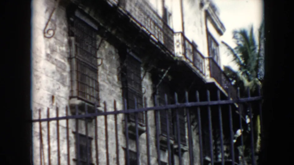 1956 Cuba frame looking past a dark iron fence toward barred windows, worn masonry and palm fronds beside the building.