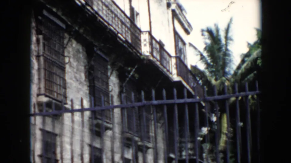 1956 Cuba home-movie frame with an angled view of barred windows, an iron gate, weathered walls and tropical planting.