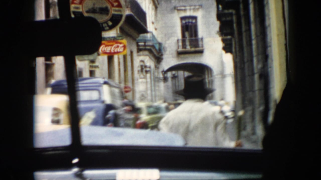 1956 Cuba home-movie frame from inside a car showing a man in a dark hat, a blue van, street traffic, and a Coca-Cola sign.