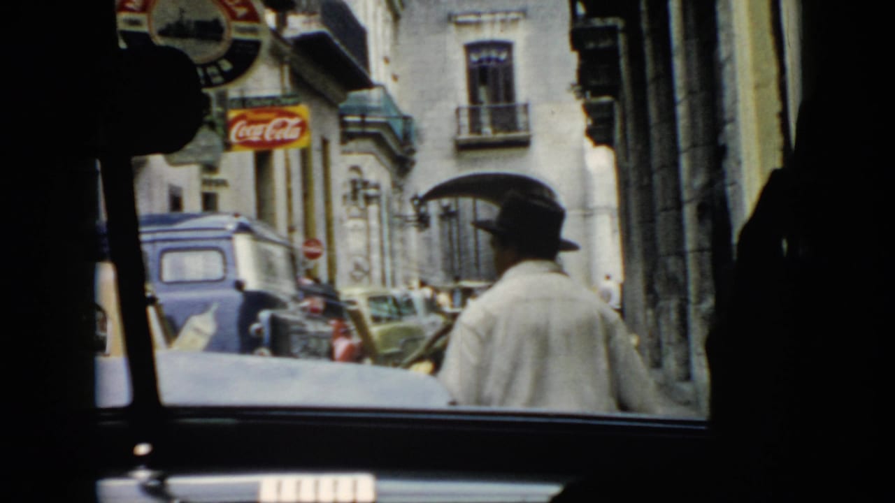1956 Cuba street view through a car window with a man in a dark hat, mid-century vehicles, building facades, and visible advertising.