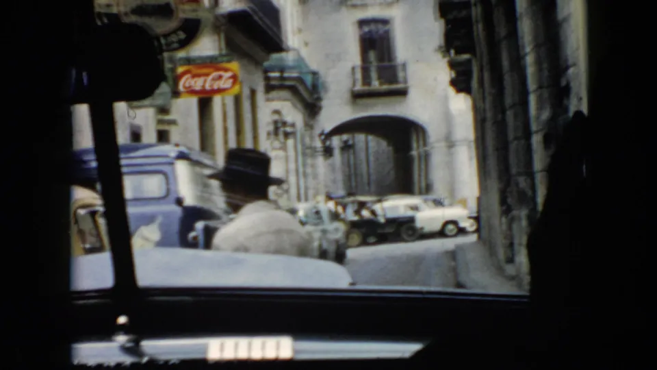 1956 Cuba passenger-view frame showing a man in a dark hat near a blue van, parked or moving cars, and advertising on buildings.