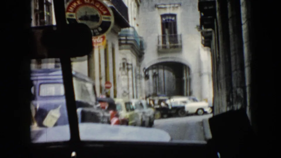 1956 Cuba home-movie frame from inside a car showing a blue van, white sedan, urban facades, signage, and fewer pedestrians in view.