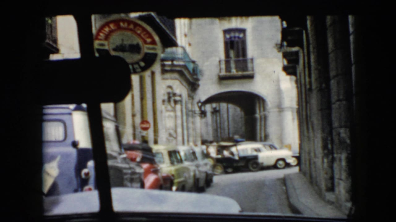 View through a vehicle toward a narrow street with parked cars, arches, and a circular sign.