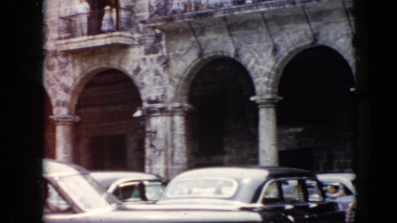 1956 Cuba street view with a stone arcade, balconies, and several parked mid-century cars in front of the building.