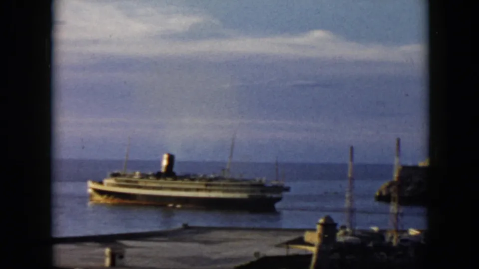 1956 Cuba home-movie frame showing a passenger steamship moving leftward through a harbor, with pavement and masts below.