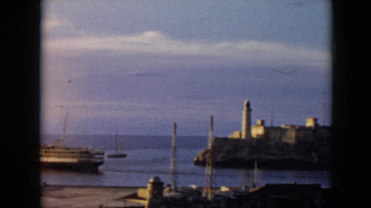 1956 Cuba home-movie frame looking through a window toward water, a passenger ship, small boats, and a lighthouse on a rocky island.