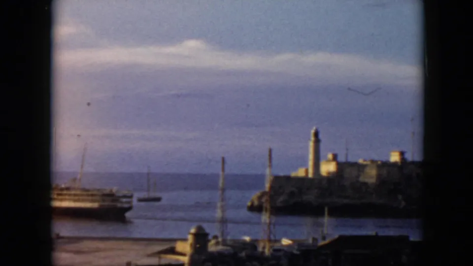 1956 Cuba home-movie frame looking through a window toward water, a passenger ship, small boats, and a lighthouse on a rocky island.
