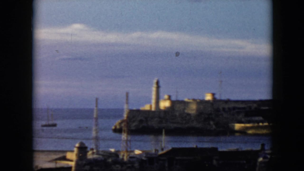 1956 Cuba travel footage frame with a lighthouse and fort-like coastal structure across the water, seen through a window opening.