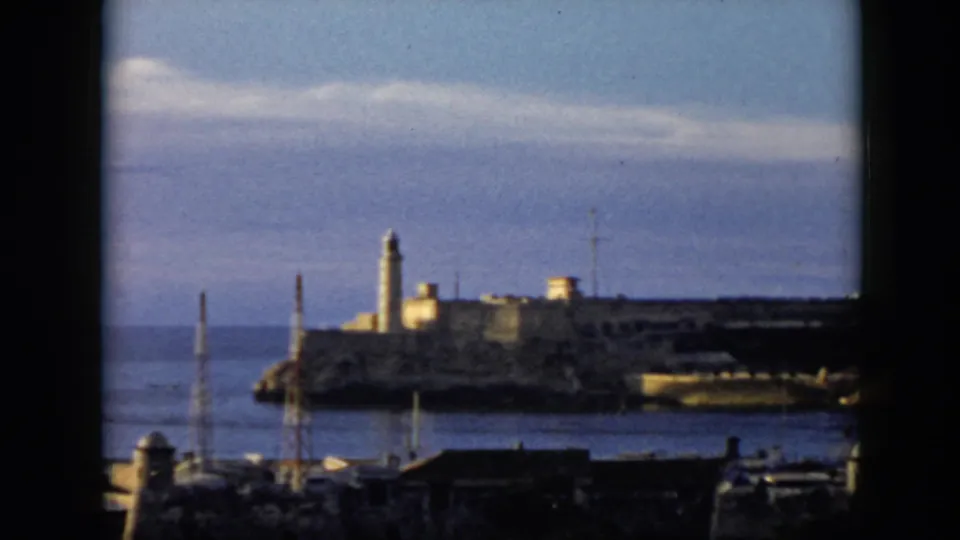 1956 Cuba 8mm frame showing a lighthouse and coastal structure across the harbor, with boat masts and dark foreground buildings.