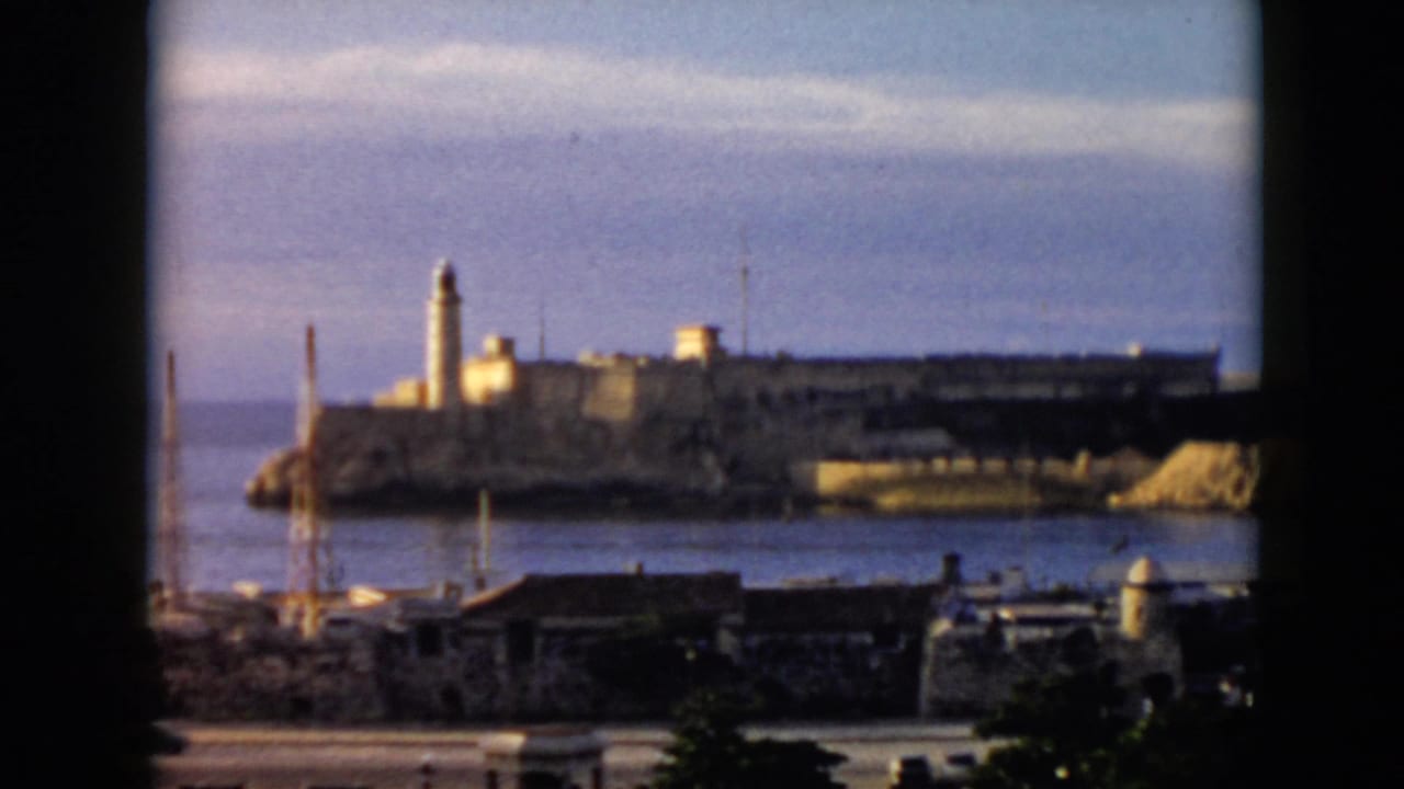 1956 Cuba home-movie frame showing Morro Castle across the water with a road and harbor structures in the foreground.