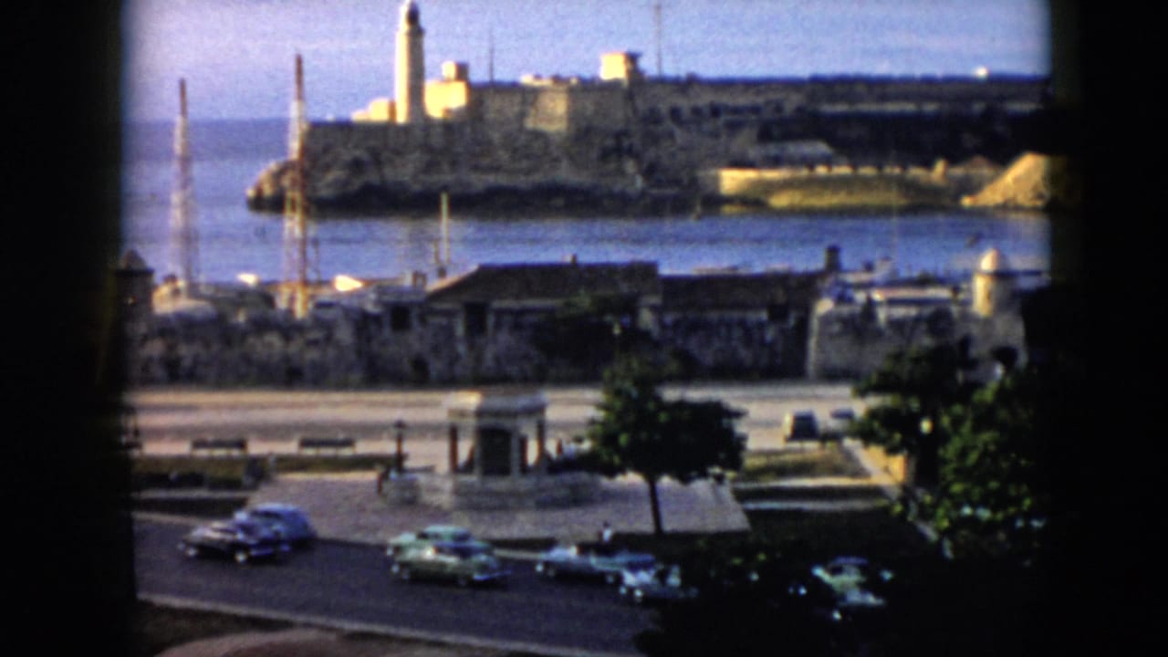 Cars move along a waterfront road with fortress and lighthouse across the harbor.