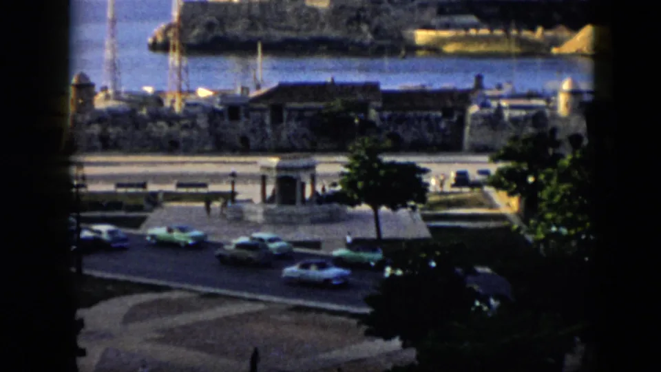 1956 Cuba street scene near Havana harbor, with classic cars, pedestrians, masts, and stone waterfront structures.