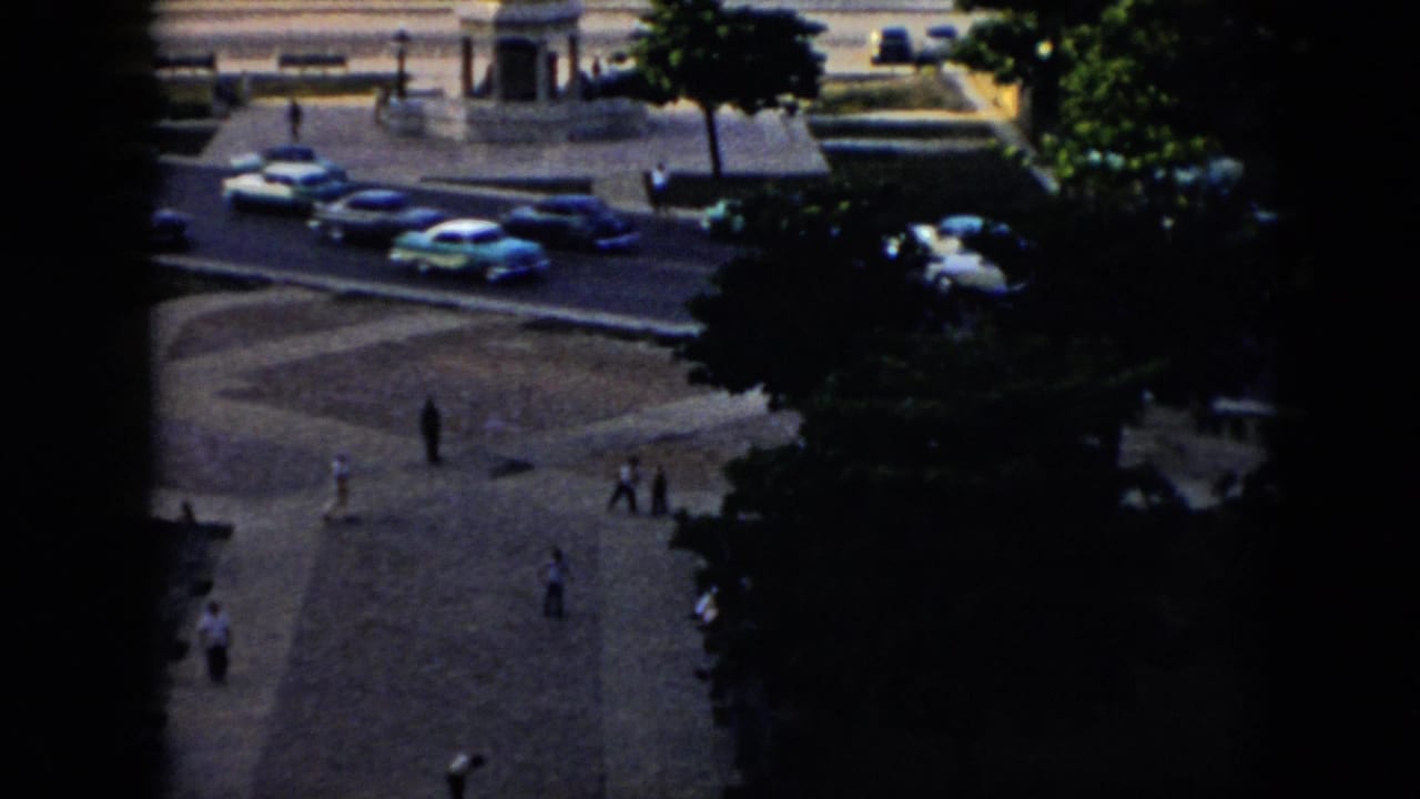 Elevated 1956 Cuba home-movie view of a public plaza with pedestrians crossing, mid-century cars on the bordering road, trees, and part of a fountain.