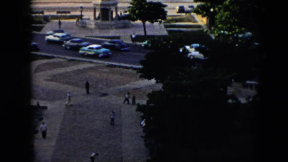 Elevated 1956 Cuba home-movie view of a public plaza with pedestrians crossing, mid-century cars on the bordering road, trees, and part of a fountain.