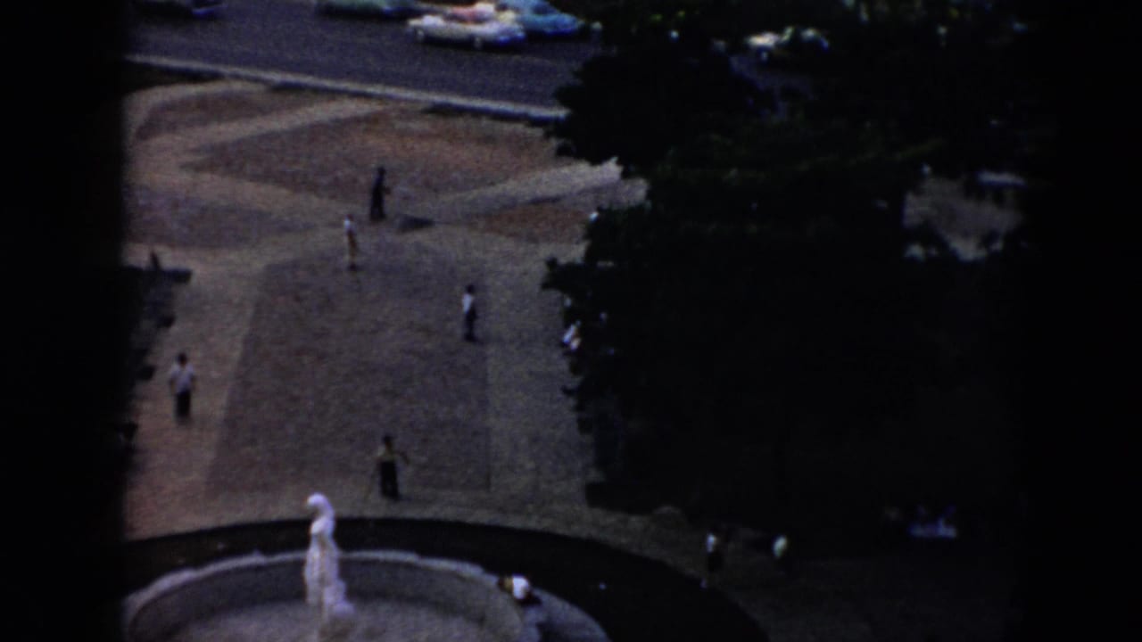 Elevated 1956 Cuba plaza view with scattered pedestrians, a circular fountain with a pale statue, shade trees, and cars on the road above.