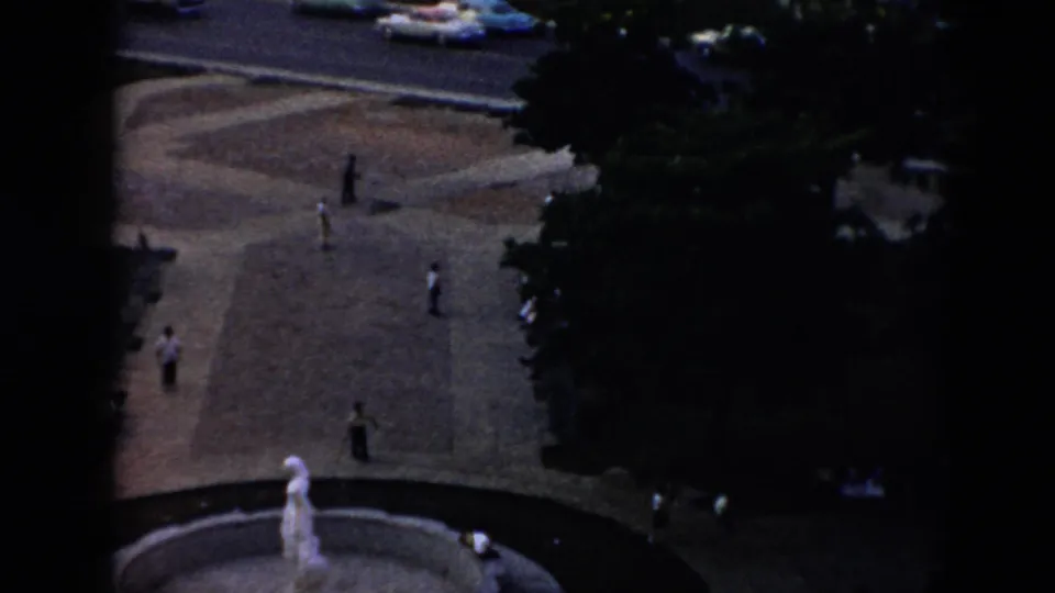 Elevated 1956 Cuba plaza view with scattered pedestrians, a circular fountain with a pale statue, shade trees, and cars on the road above.