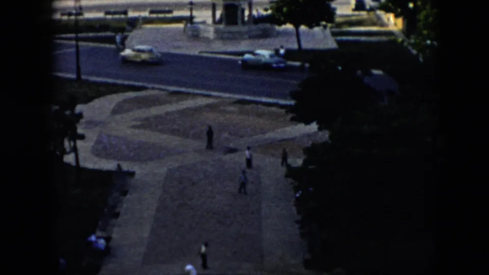 Elevated 1956 Cuba street-and-plaza view with mid-century cars, pedestrians, dense trees, patterned paving, and the fountain statue near the lower edge.