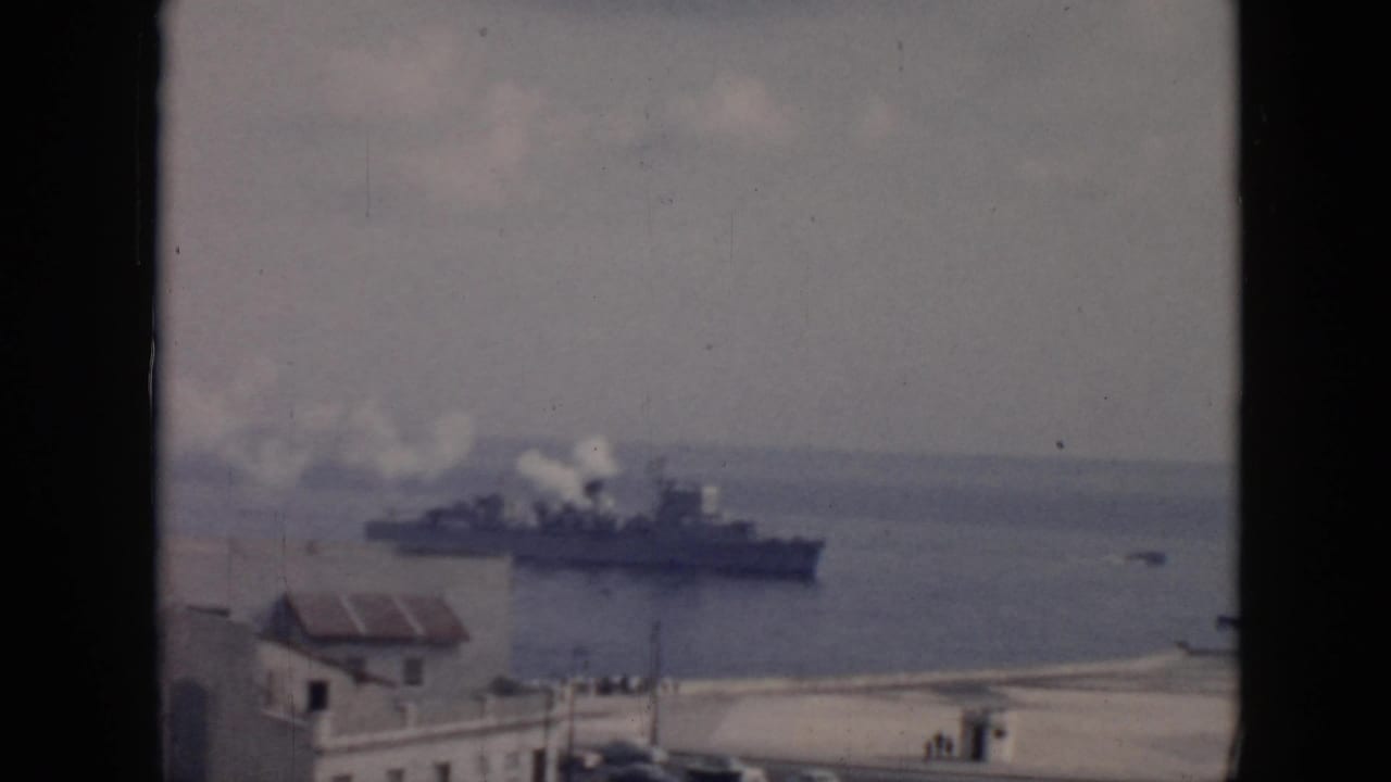 1956 Cuba harbor view with a large military-style ship smoking offshore and low coastal buildings in the foreground.