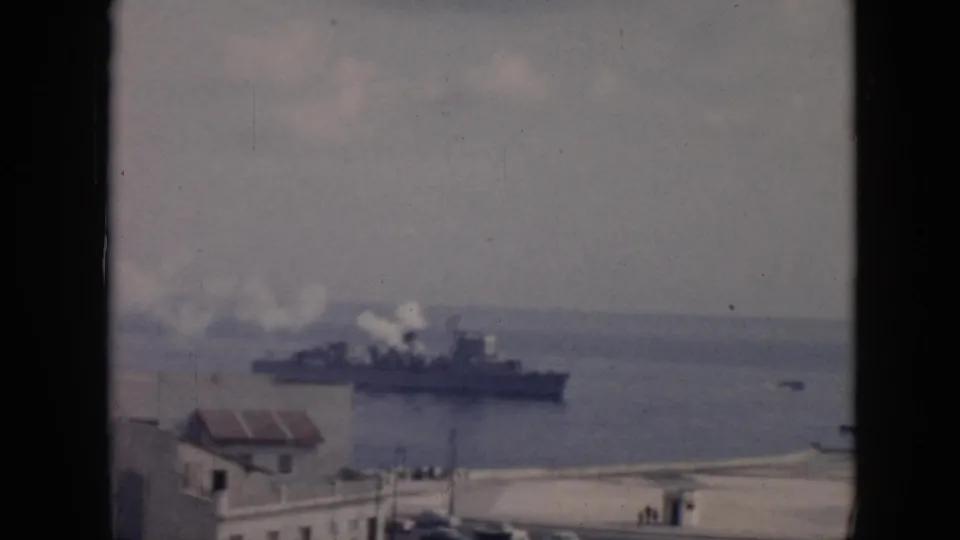 1956 Cuba harbor view with a large military-style ship smoking offshore and low coastal buildings in the foreground.