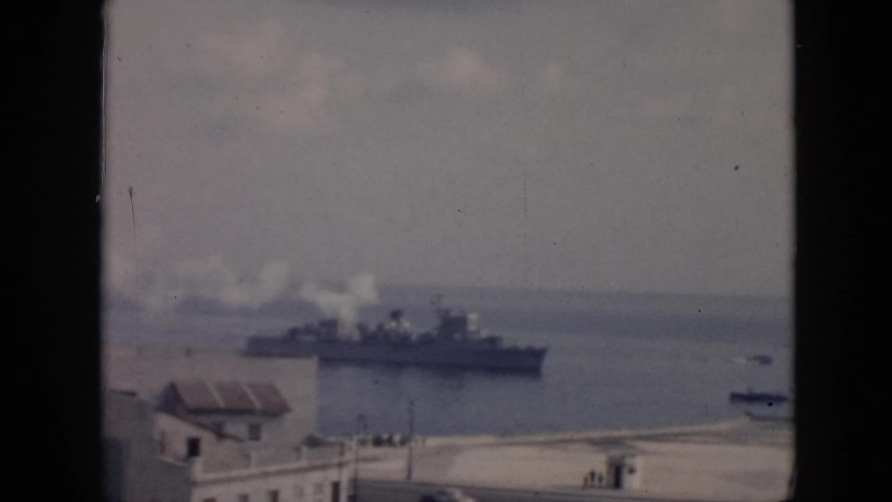 A large vessel with white smoke from its stacks crosses a Cuba harbor, seen from an elevated shoreline viewpoint.