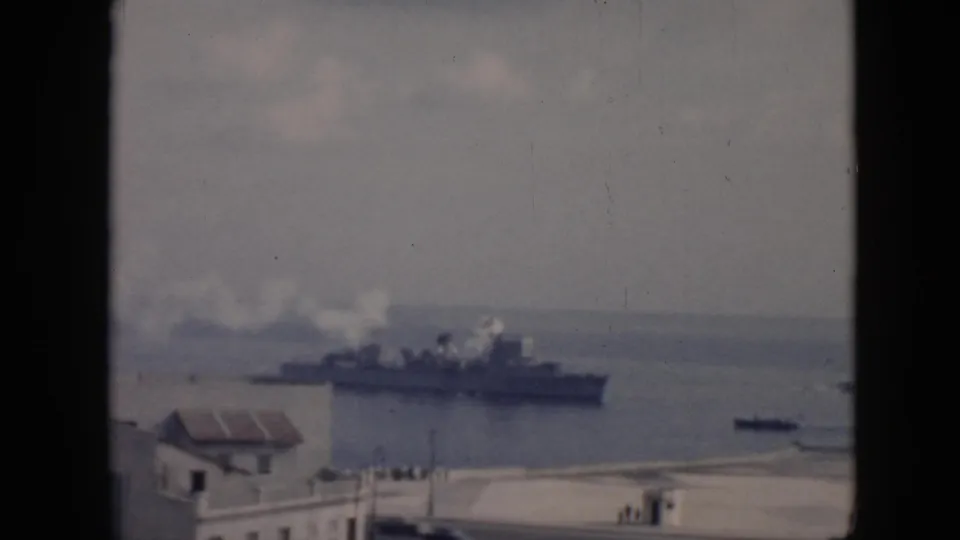 Elevated 1956 Cuba waterfront view showing a smoking military-style ship beyond a low building and small harbor boat.