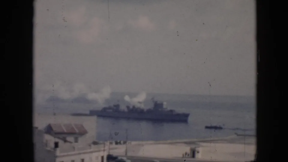 The military-style ship remains offshore as white smoke trails above the harbor and small shoreline details appear below.
