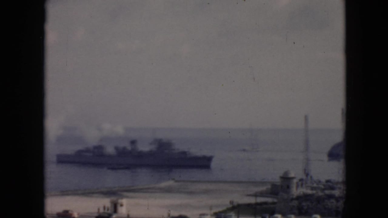 1956 Cuba 8mm frame showing a large naval-style ship offshore, smoke near its stacks, and a coastal town foreground.