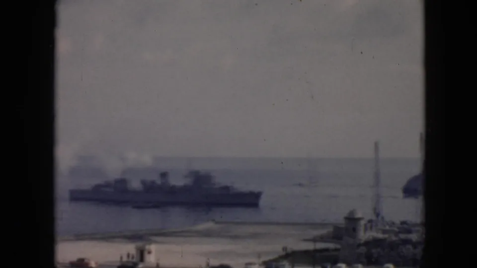 1956 Cuba 8mm frame showing a large naval-style ship offshore, smoke near its stacks, and a coastal town foreground.