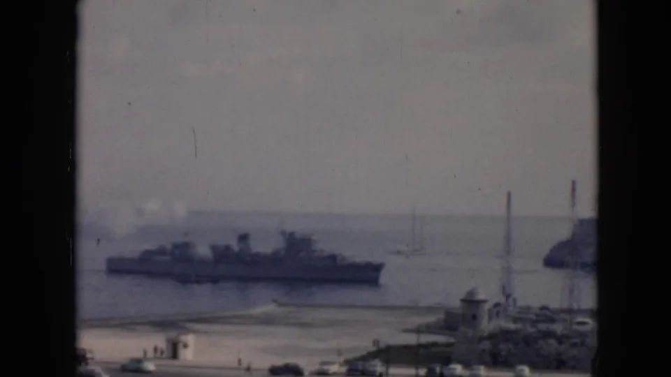 1956 Cuba 8mm frame showing a large ship in the harbor, shoreline structures, a dark vehicle, and distant water traffic.
