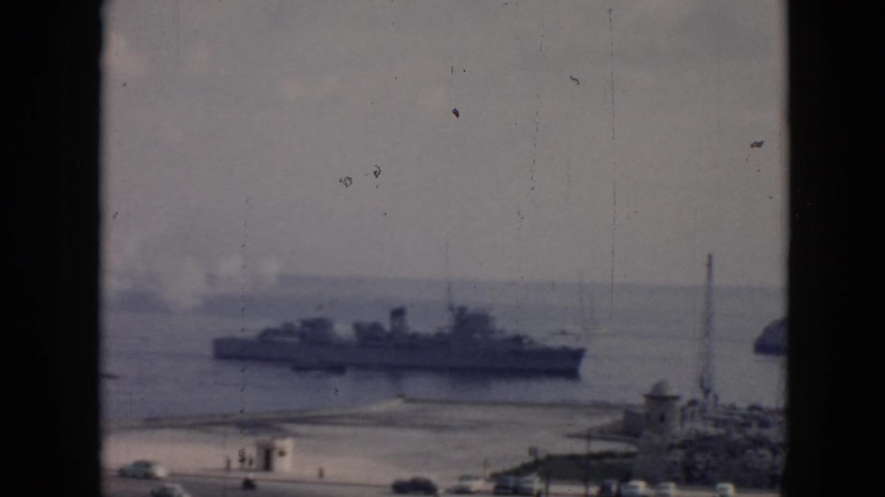 1956 Cuba home-movie frame showing a large harbor ship seen through a dark window frame, with shoreline activity below.
