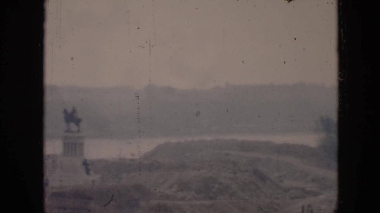 1956 Cuba home-movie frame with a distant mounted statue, water, rolling land, and a dark vertical window divider.