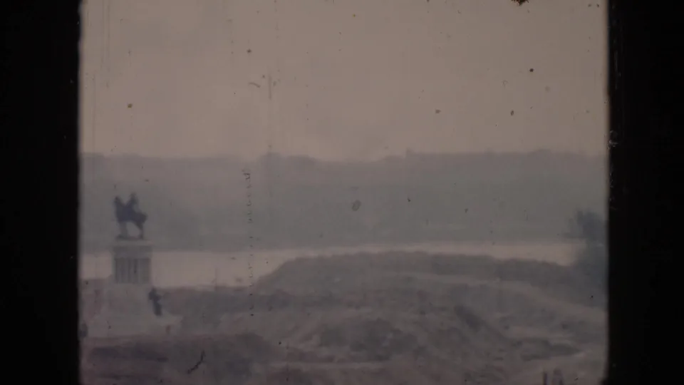 1956 Cuba home-movie frame with a distant mounted statue, water, rolling land, and a dark vertical window divider.