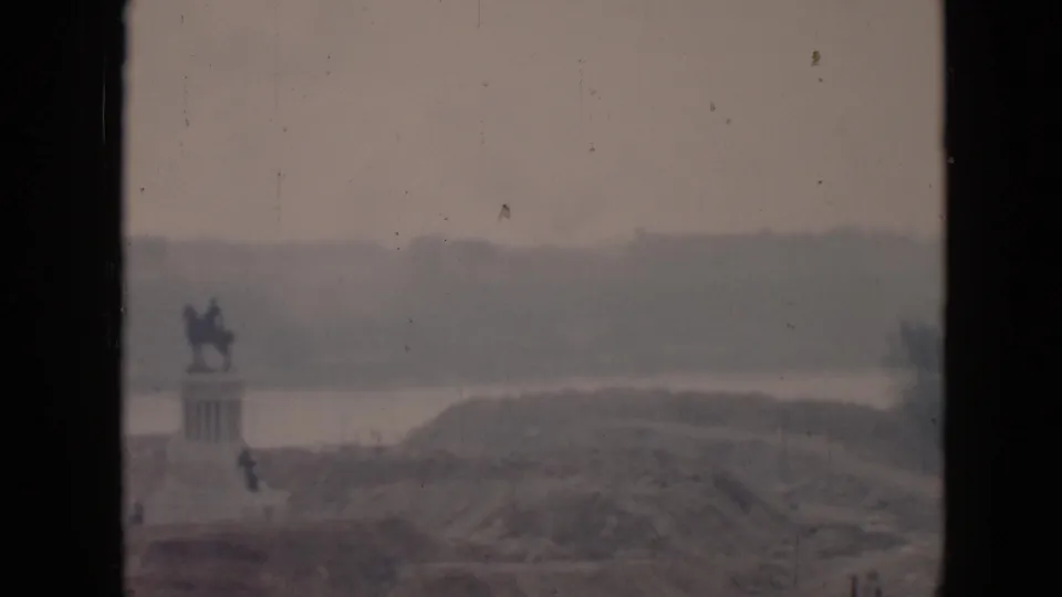 1956 Cuba 8mm still showing a distant equestrian monument, a pale water band, and soft hills beyond the foreground terrain.