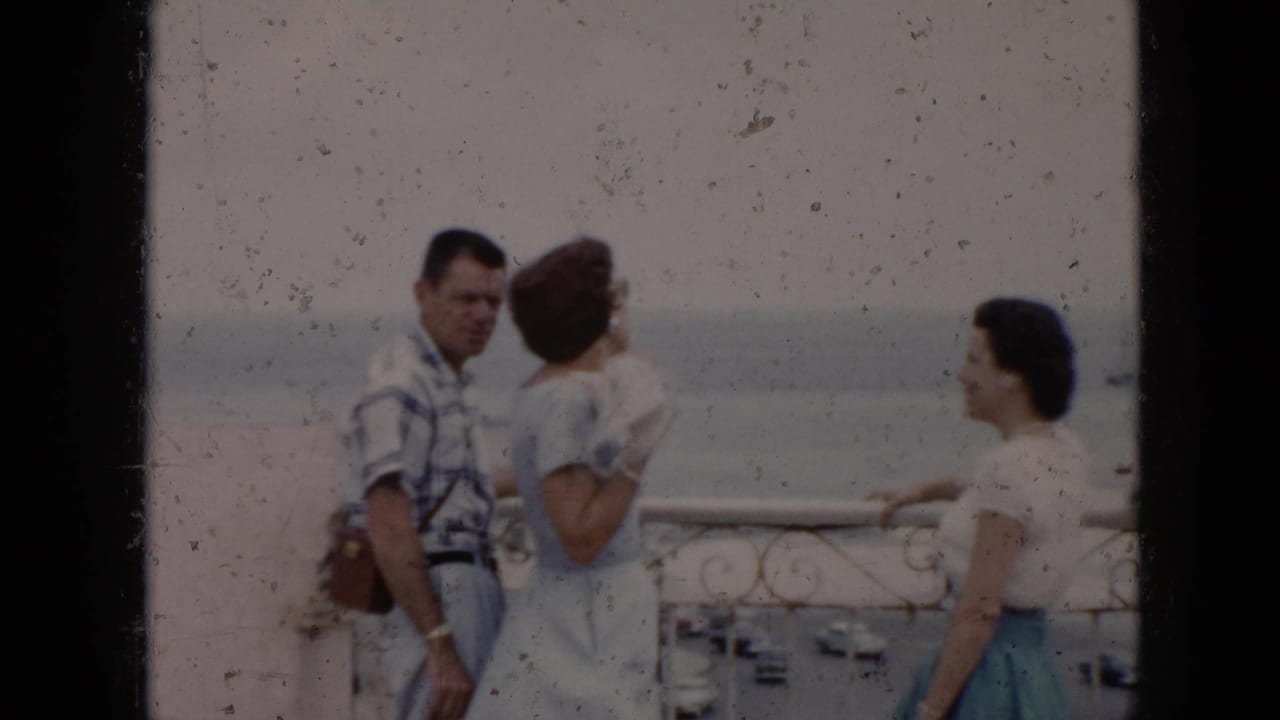 Three adults stand by a balcony railing overlooking water and a street below.