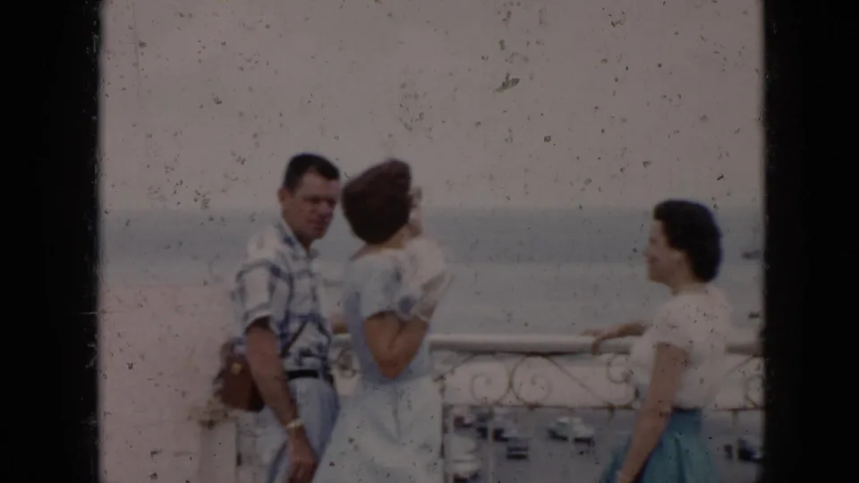 In 1956 Cuba, a man in a plaid shirt and two women stand at a balcony railing with water and traffic visible below.