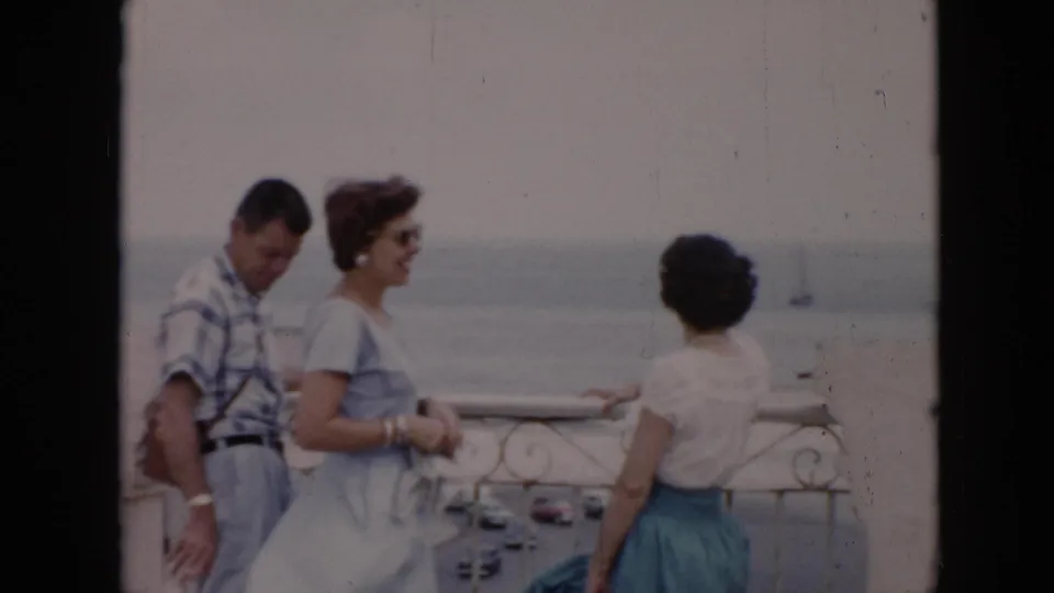 Three adults in mid-century clothing stand on a Cuba balcony, with a coastal street, cars, water, and a small sailboat in the distance.