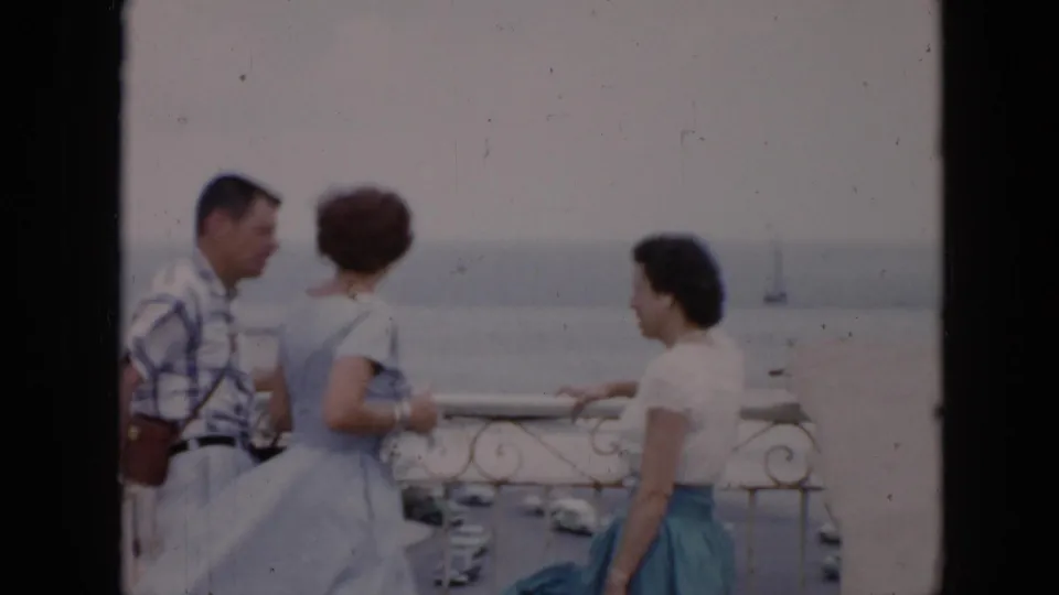 A 1956 Cuba balcony moment shows three adults conversing beside a railing, with a waterfront street, cars, and sailboat behind them.