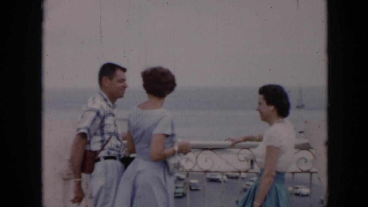 A man and two women talk beside a railing with the sea behind them.