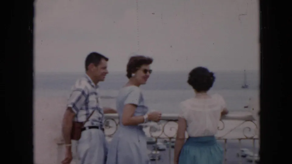 A woman in a light blue dress looks toward the camera as the group stands at a Cuban coastal overlook in soft 8mm color.