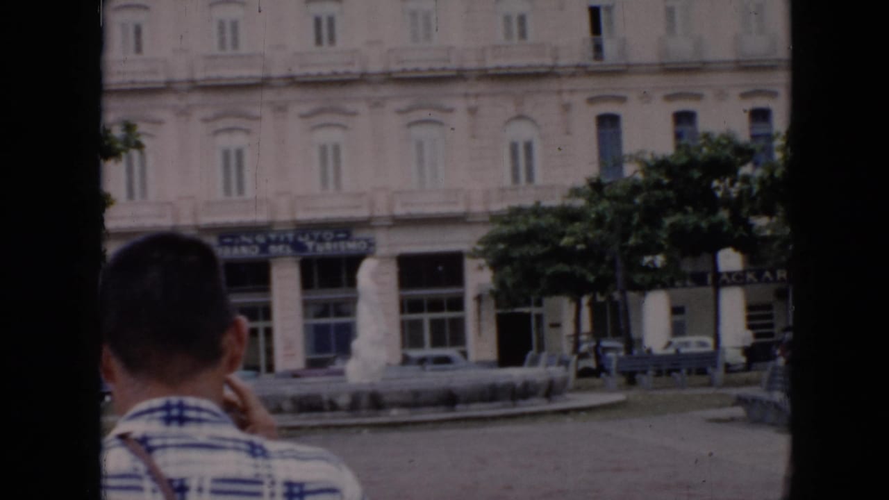 1956 Cuba home-movie frame showing a foreground visitor, a white city-center building, tourism signage, trees, cars, and a fountain.
