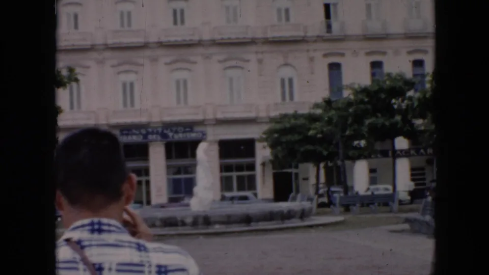 1956 Cuba home-movie frame showing a foreground visitor, a white city-center building, tourism signage, trees, cars, and a fountain.