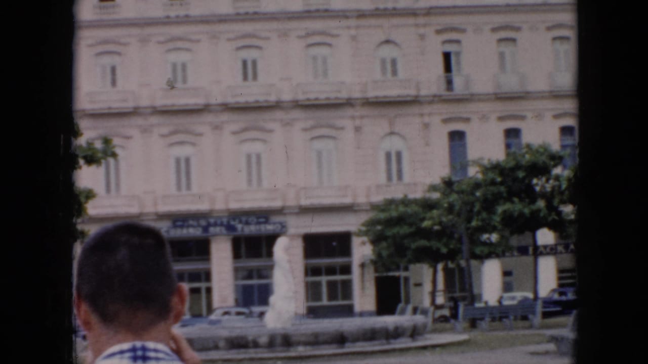 White multi-story building with balconies, storefront signs, trees, cars, and a man at the lower edge.