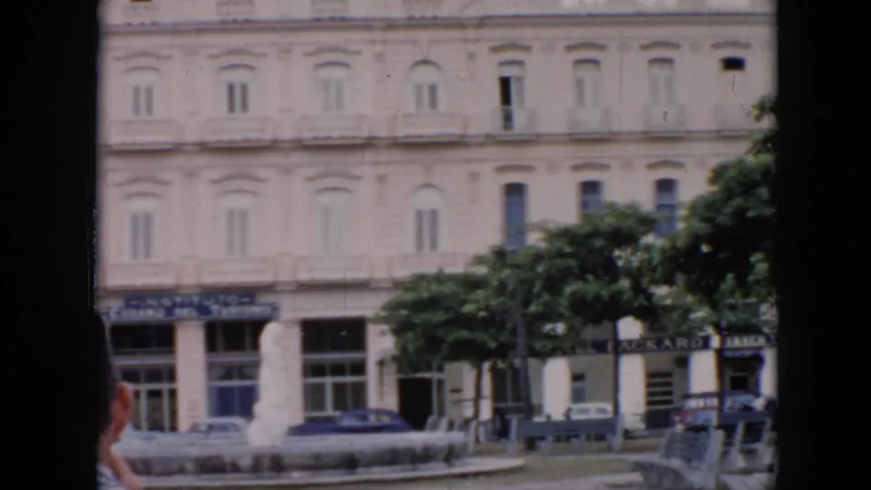 1956 Cuba home-movie frame of a city building, street trees, fountain base, parked cars, and partial foreground visitor.