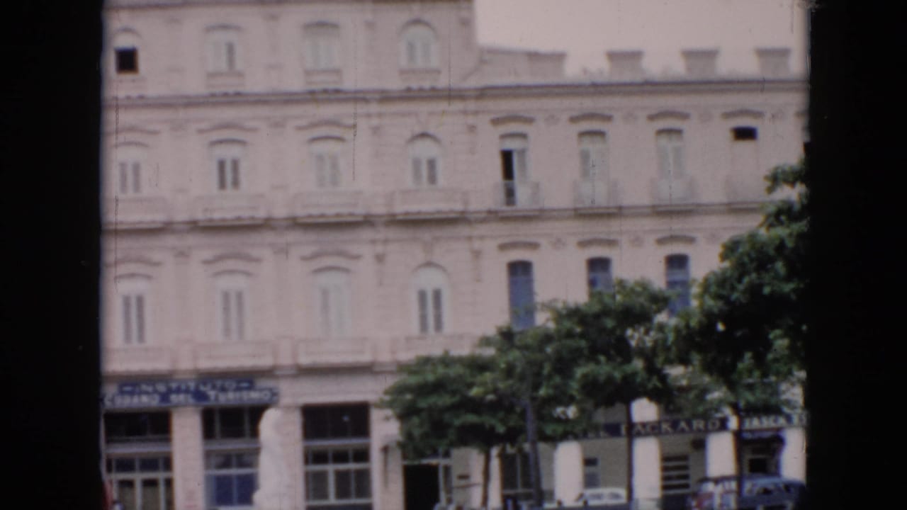 1956 Cuba 8mm frame showing a pale classical building facade, leafy trees, and street-level signage partly obscured by dark foreground edges.